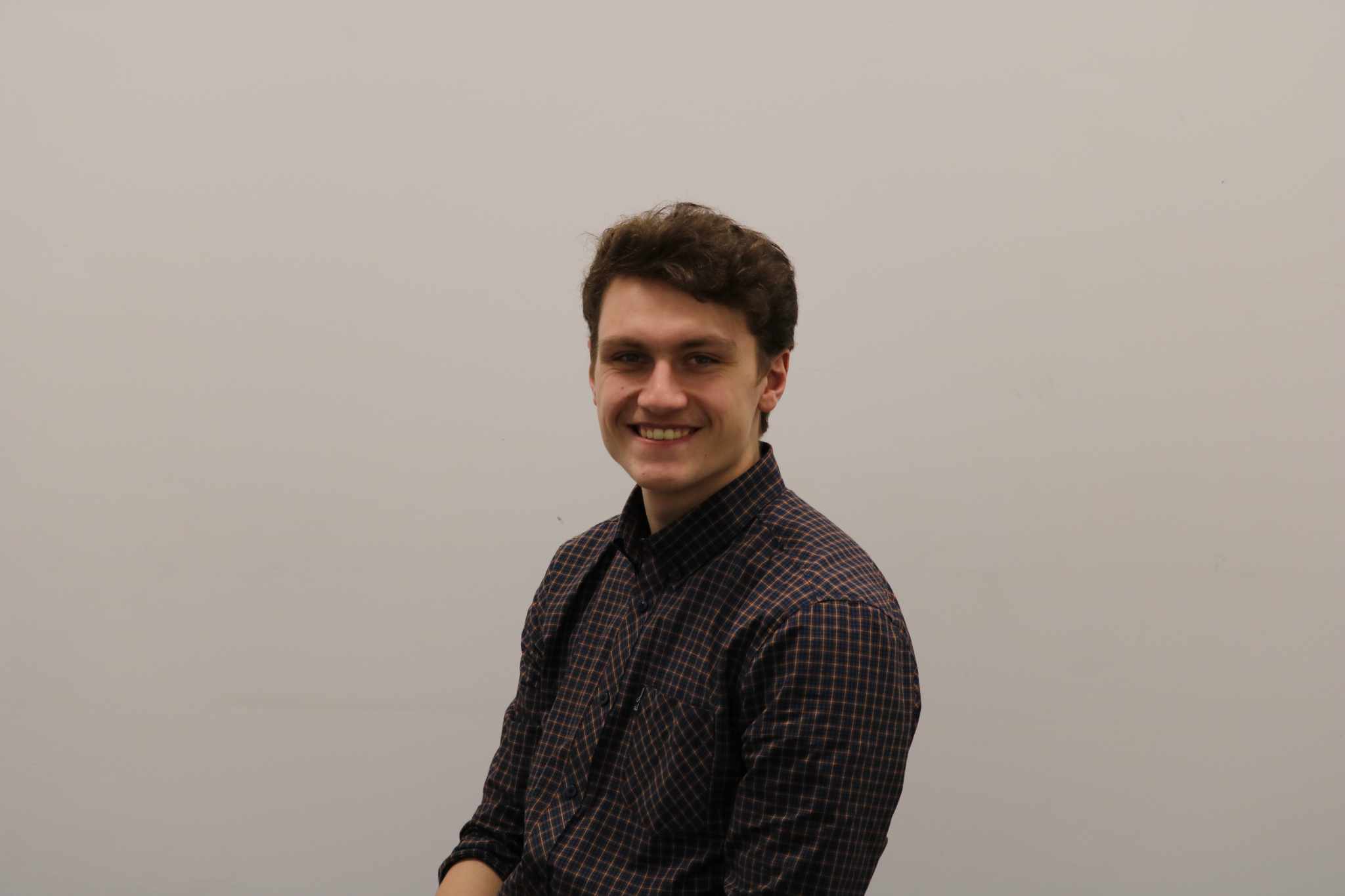 A headshot of Jonah Davies-Jones in a checkered shirt smiling
                against a light grey background, with his body turned slightly
                to the left.